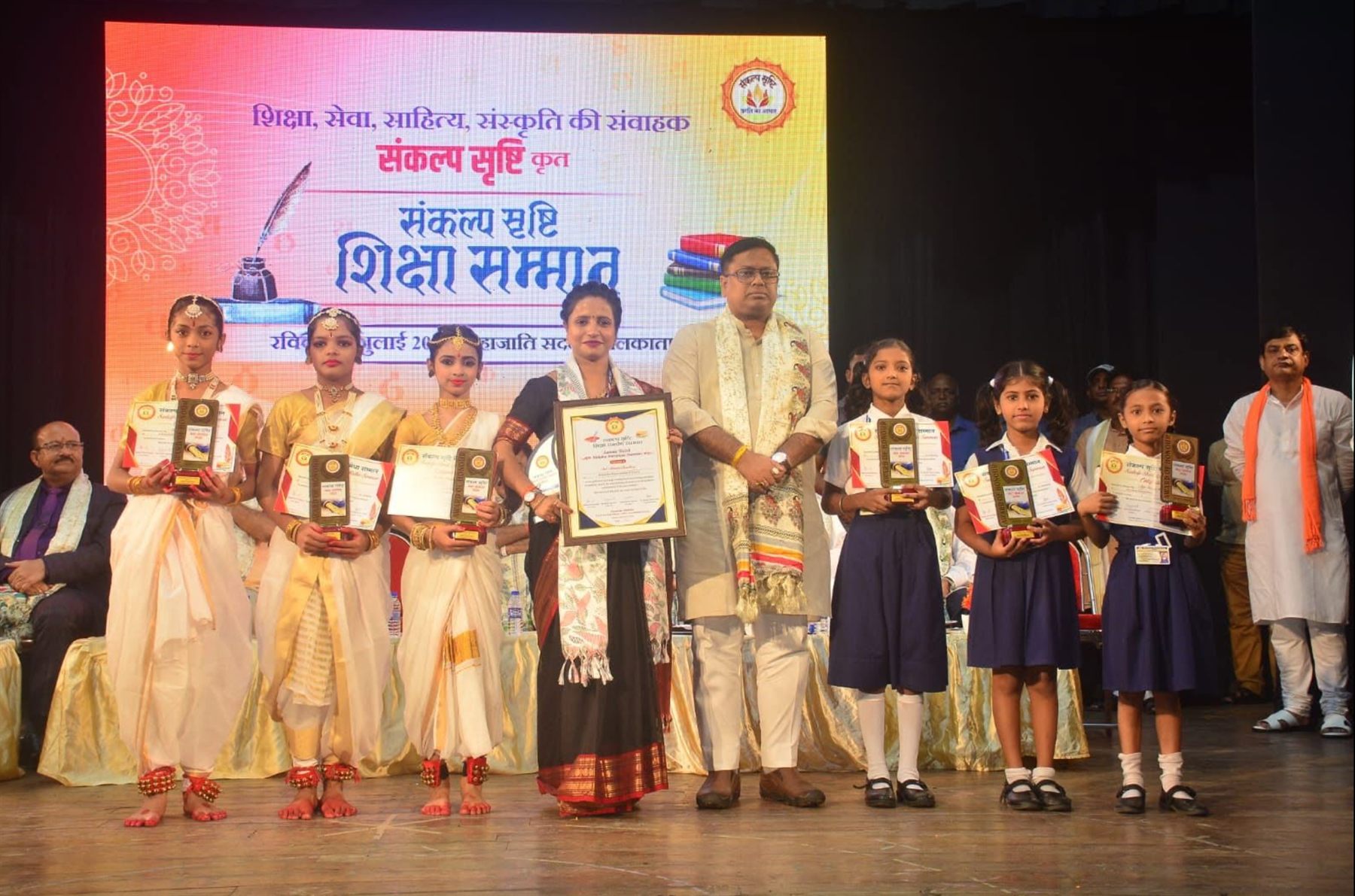 Students of Arya Kanya Mahavidyalaya on stage at Mahajati Sadan, Kolkata, holding certificates and trophies at the Sankalp Srishti Shiksha Samman ceremony.